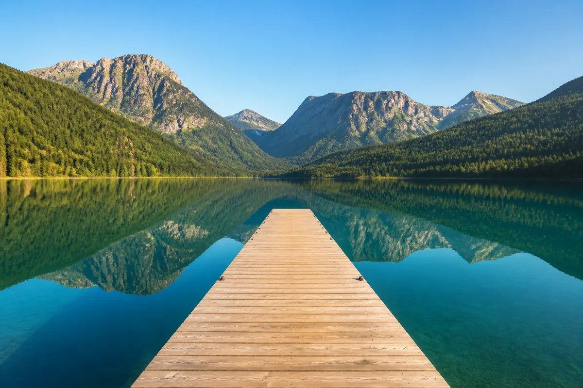 Ponton de bois avançant sur une eau turquoise parfaitement calme, montagnes en arrière-plan à la lumière de fin de journée