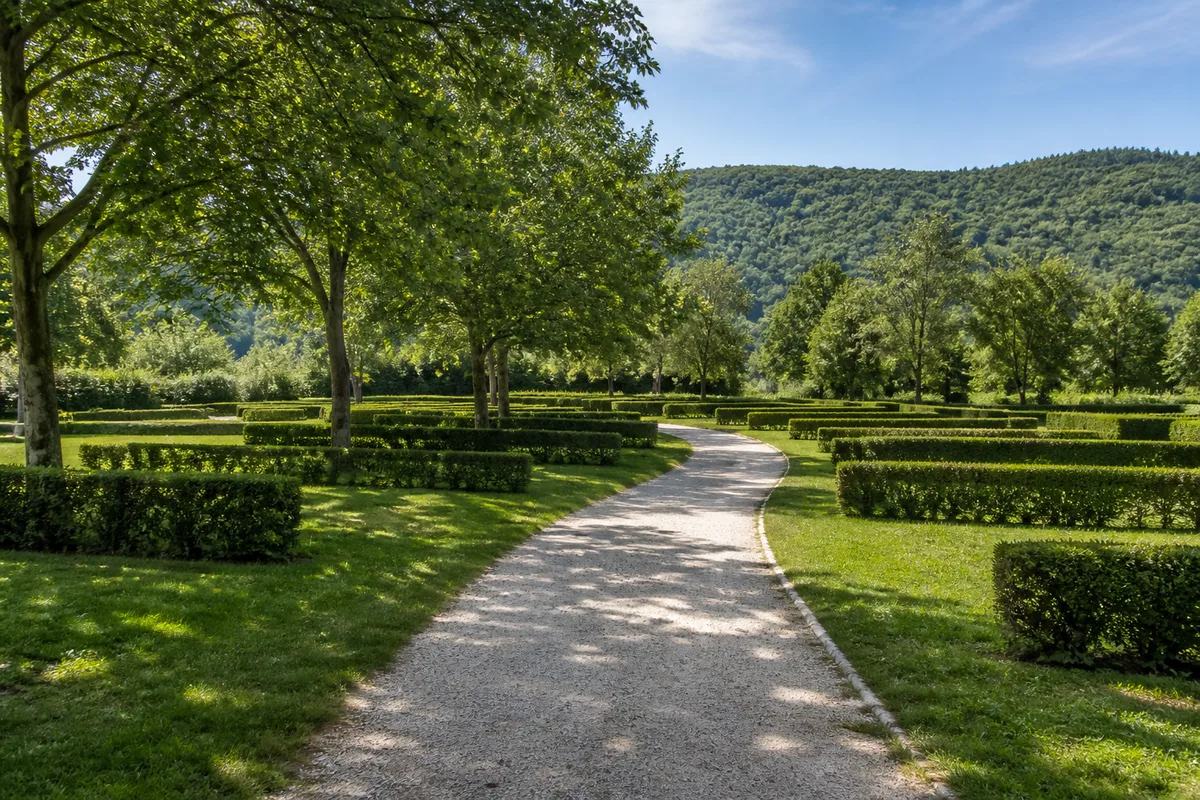 Chemin de gravier clair traversant un terrain de camping désert, emplacements herbeux vides bordés de haies basses
